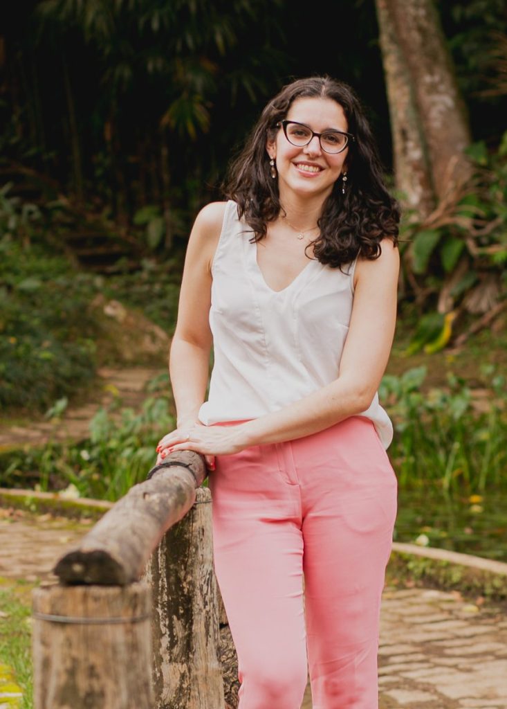 Dra Fernanda Belem em pé, com óculos, uma clusa branca e calça rosa num parque em cima de uma ponte de madeira