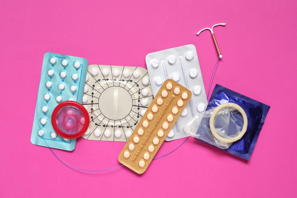 imagem de métodos contraceptivos como algumas cartelas de comprimidos, diu e preservativos em fundo rosa