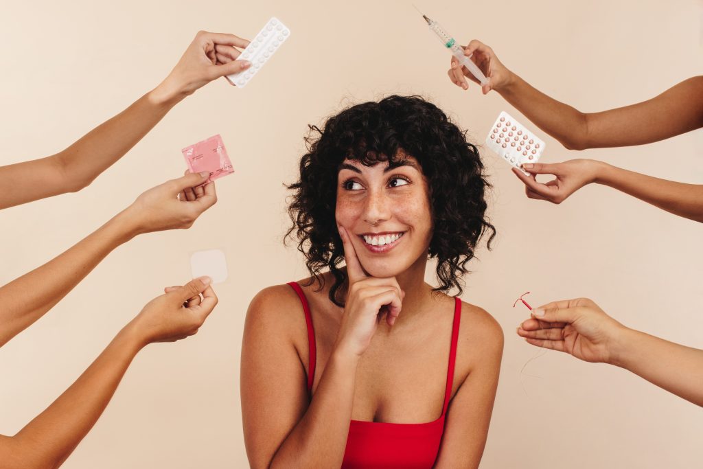 Mulher de cabelo castannho médio e camiseta vermelha com a mão no queixo sorrindo olhando para alguns métodos contraceptivos expostos, sendo segurados por algumas pessoas